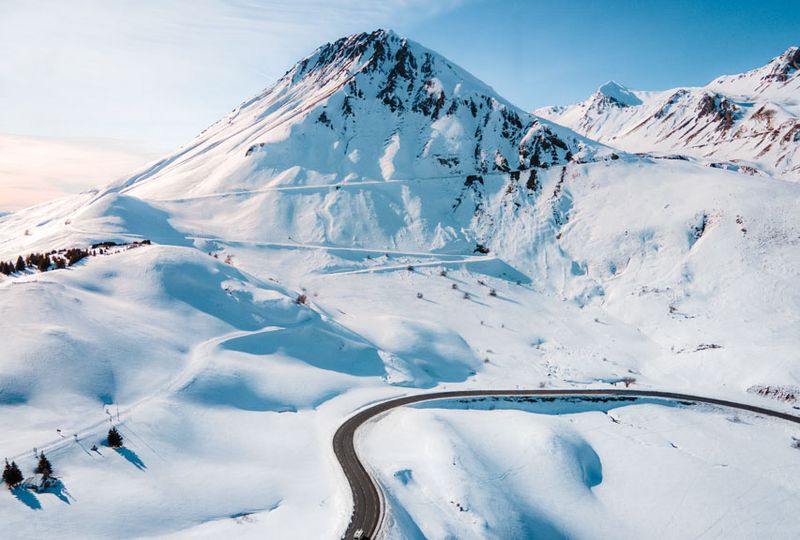 A drone shot of a winding mountain road, surrounded by fresh snow and a high mountain peak