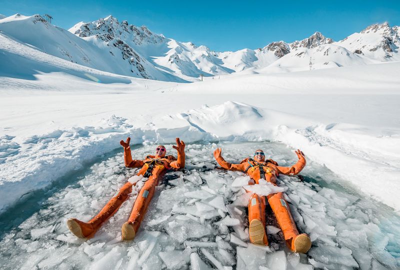 Two people dressed in orange suits floating in an icy lake, giving a thumbs up to camera. Snow surrounds the hole broken in ice and snow to reveal the water