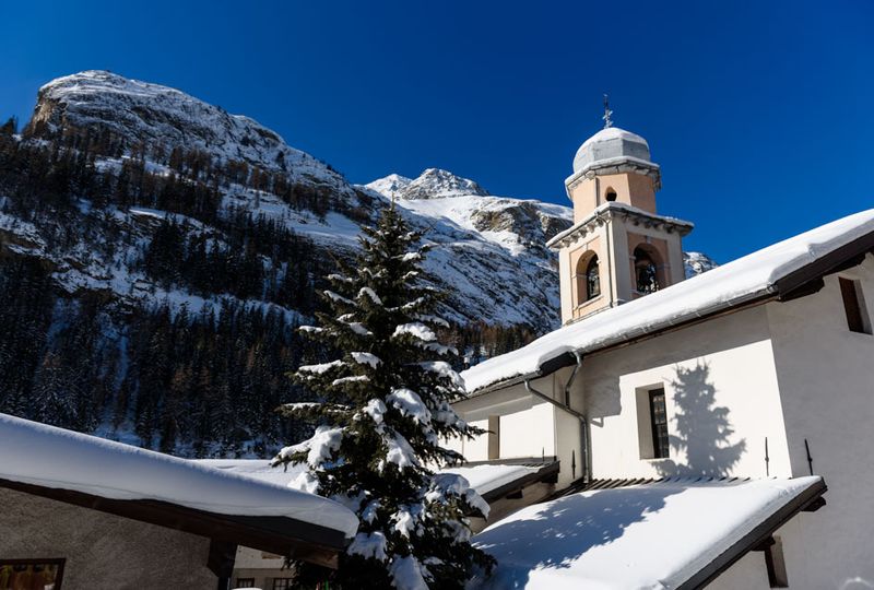A picture of Tignes' church under snow, with blue sky, snowy mountains and a pine tree in shot