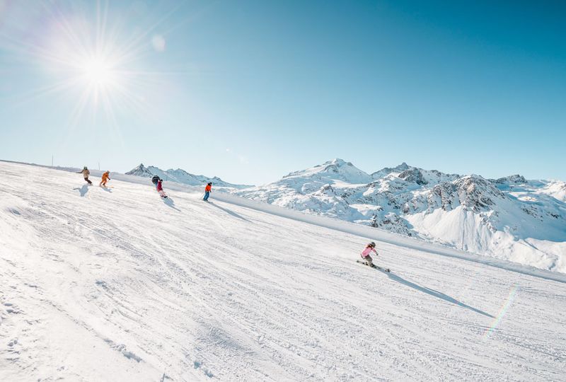 a group of skiers make their way down a wide piste, snowy mountain tops in the background suggest they're high up skiing in the resort of Tignes