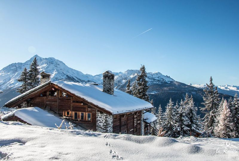 A big wood and stone chalet is pictured, with snow on the roof, in front of a grand view across snowy mountains covered with trees in background