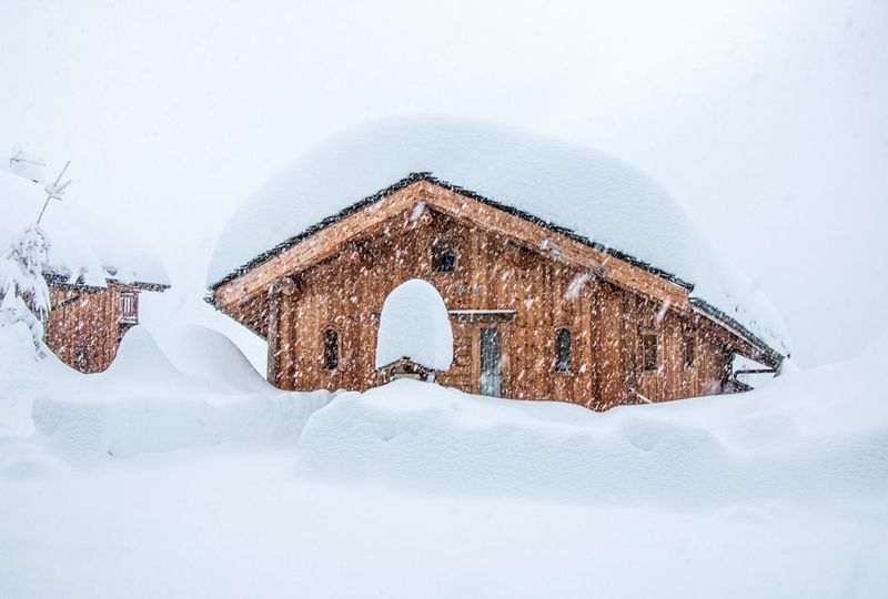 A wooden hut sits with a few feet of fresh snow piled on the rood, surrounded by fresh, white snowfall