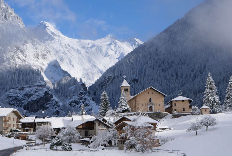 A snowy alpine village sits in a high valley, surrounded by snowy peaks and trees. The church steeple is covered with snow