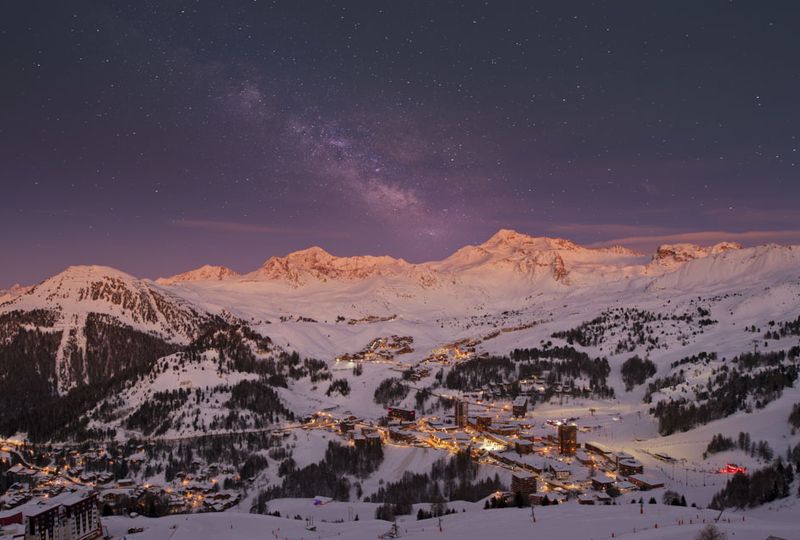 A night time shot of a snowy alpine village, lit up by lights, with even the snowy peaks in the far distance glowing orange under the night's sky