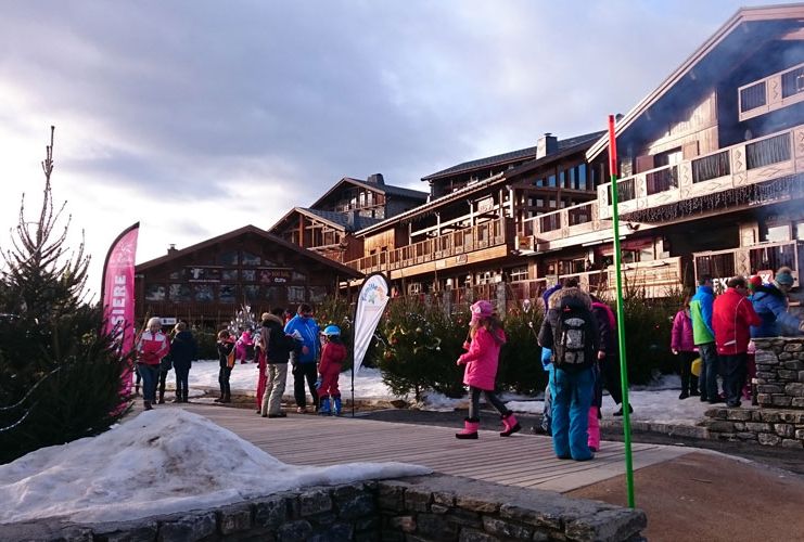 Winter tourists walk along a pavement, with a little snow piled to the side, in front of big wooden chalet buildings