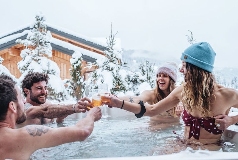 four friends in a hot tub cheersing with drinnks - very snowy setting around them