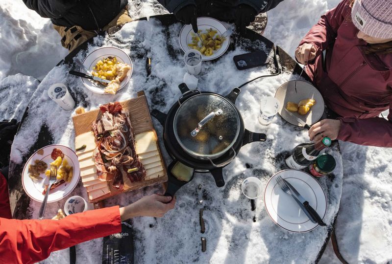 A table is dusted with snow and covered with alpine food - a meats and cheese platter and a pan with a lid