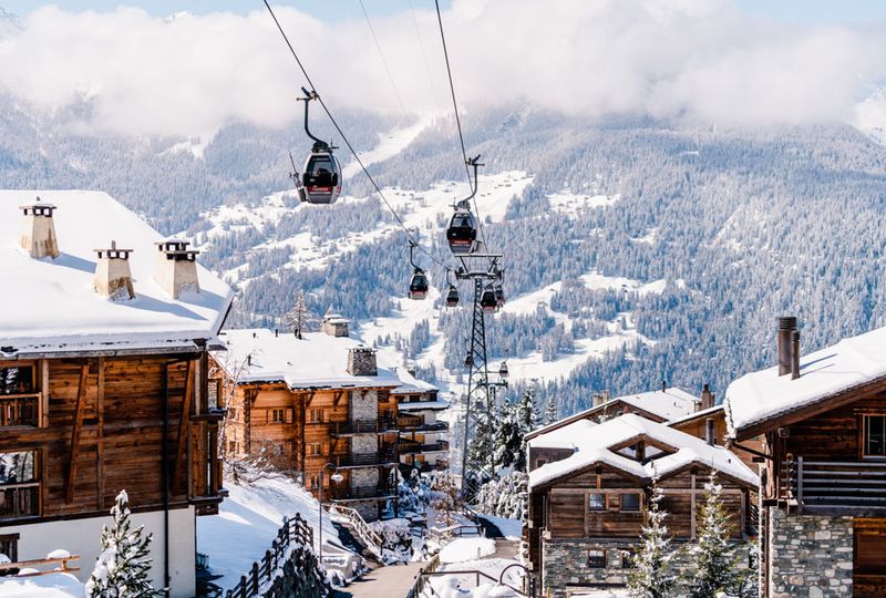 A gondola travels over the wooden buildings of Verbier, with snowy slopes in the background, the main street below