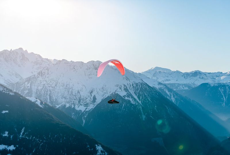 A red parapente flies over the valley, with the snow-line visible on the mountain behind