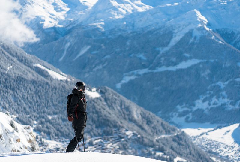 A skier looks towards the camera, standing still against a good looking backdrop of snowy trees on a facing slope across the valley