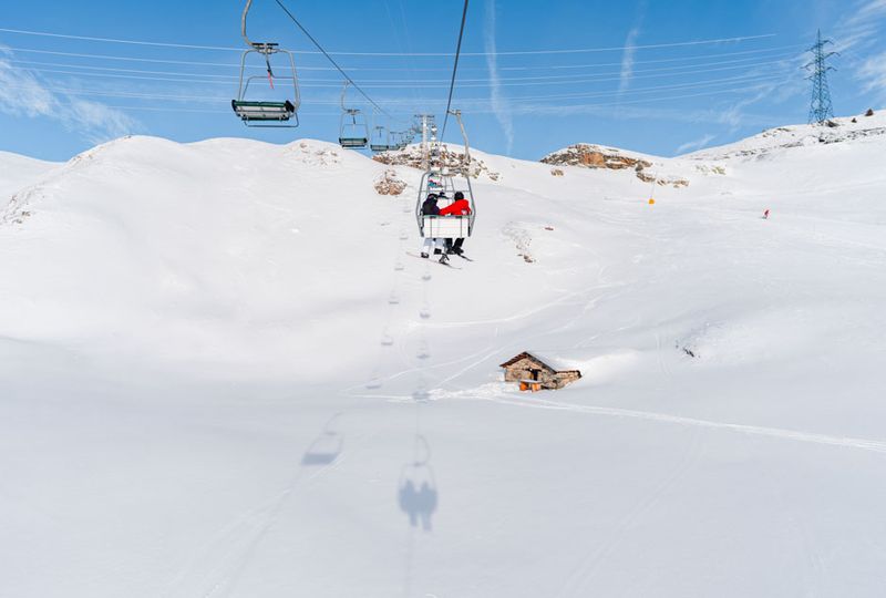 A chairlift travels over untouched snow, casting a perfect shadow beneath. Two skiers ride the chairlift