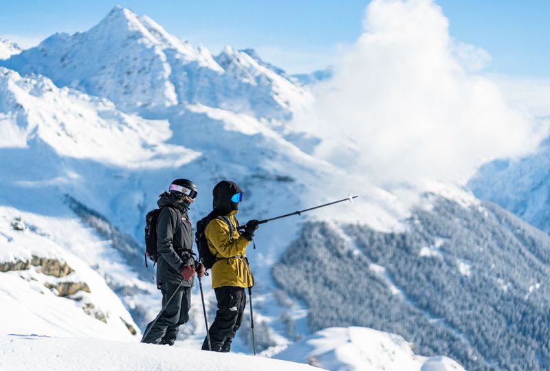 Two skiers stand shoulder to shoulder, one pointing at something in the distance out of shot. The scene is a seriously snowy one, with a tall white mountain in the distance behind skiers