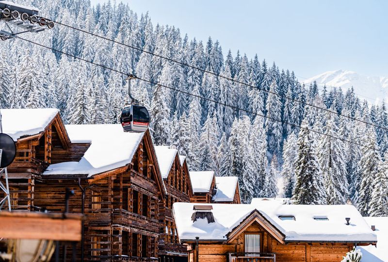 Beautiful wooden chalets with snow on roofs sit in front of snow-laden trees. A gondola travels over the roofs of the houses