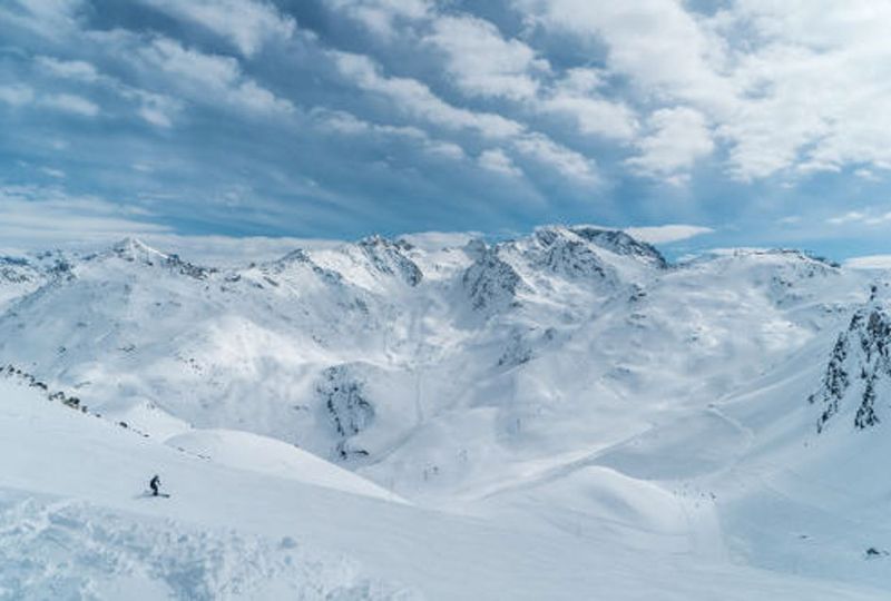 A big mountainscape shows a lone skier on a cat track, at the top of the mountain