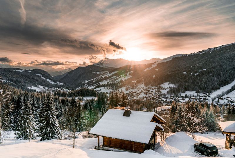 One mountain cabin has a snowy roof, shot at sundown with an orange light