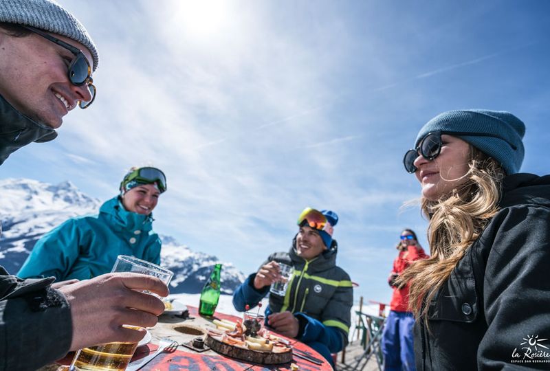 Group of adults sit around a table outside on ski slopes under blue skies drinking beer