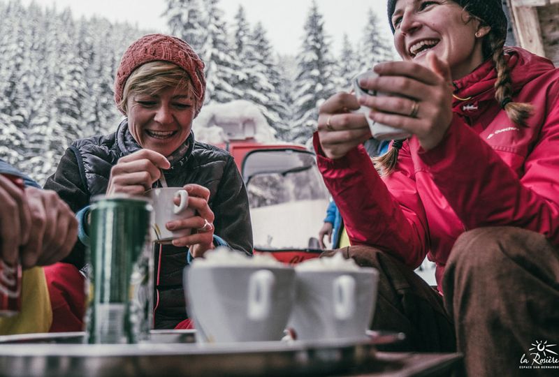 two woman hold onto hot drinks, laughing, at an outside cafe surrounded by snow