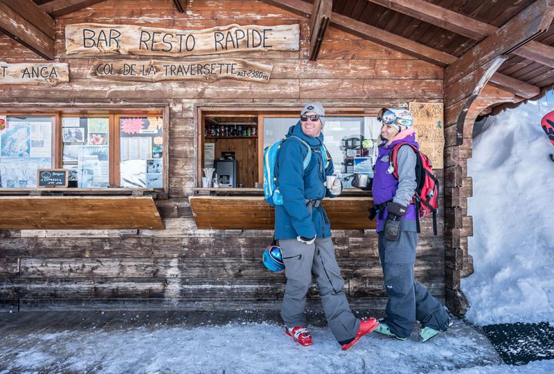 Two skiers stand at a self-service hut hatch holding hot drinks on a pit stop from skiing