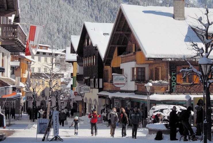 The main street of ski village St Anton during the day, with snow covered roofs, skiers carrying skis, and shoppers and walkers pictured