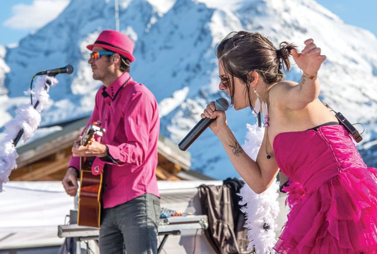 Singer in pink dress sings into microphone, her guitarist in pink plays behind, against very snowy mountain backdrop blurred in background